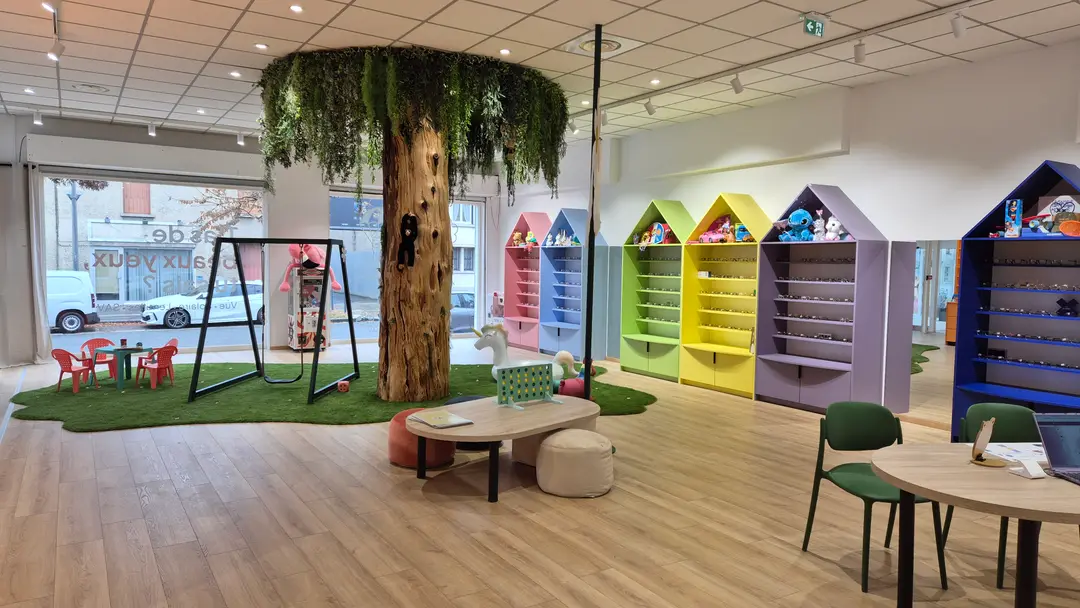Vue d'ensemble du magasin : l'arbre au centre, la balançoire, et la rangée de meubles en forme de maisons colorées où sont présentées les montures enfant.